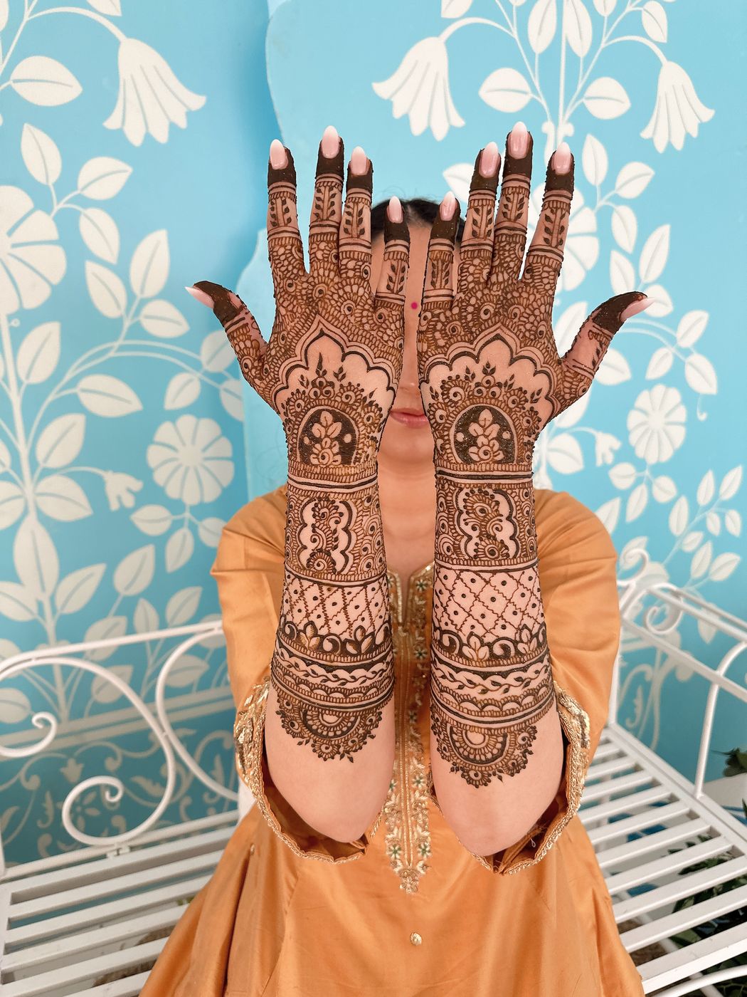 A bride holding up both hands to show full bridal henna against a floral backdrop